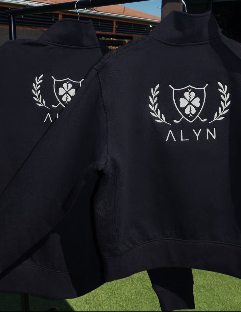 Two black quarter zips with white logos and 'ALYN' text hanging on a clothesline against a blue sky.