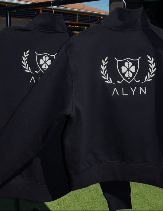 Two black quarter zips with white logos and 'ALYN' text hanging on a clothesline against a blue sky.
